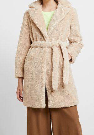 Femme portant un manteau teddy beige et duveteux avec un col large et une ceinture, associé à un pantalon marron et une chemise vert clair en dessous.