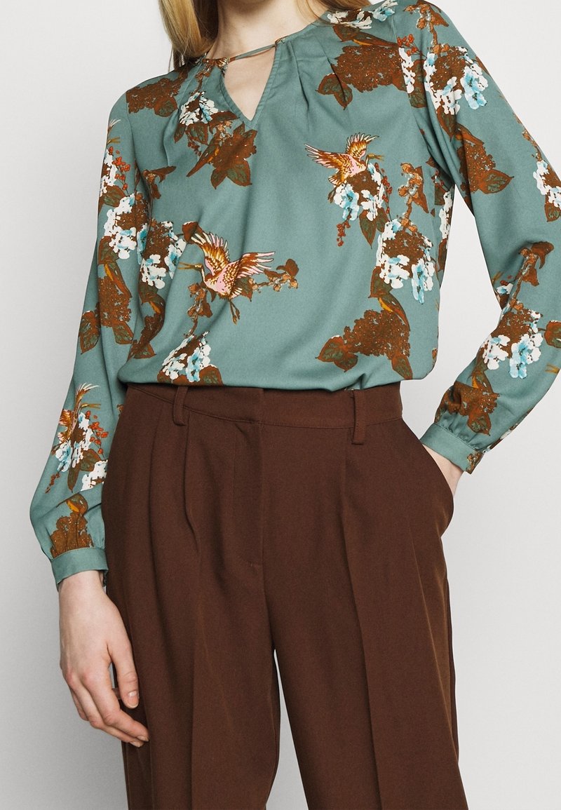 Blouse florale verte à manches longues, présentant une petite ouverture en forme de clef au cou, ornée d'oiseaux et de motifs floraux. Pantalon marron.