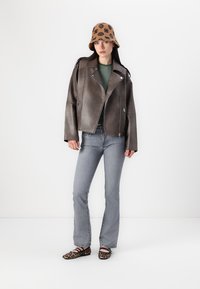 Veste de biker en cuir gris foncé avec des fermetures éclair argentées, chemise rayée verte, jean flare gris clair, et chapeau seau marron avec un motif noir.