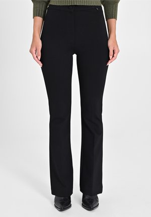 Kvinde iført sorte bukser med lige ben, kombineret med sorte spidse sko og en olivengrøn ribstrikket sweater, står mod en ensfarvet baggrund.