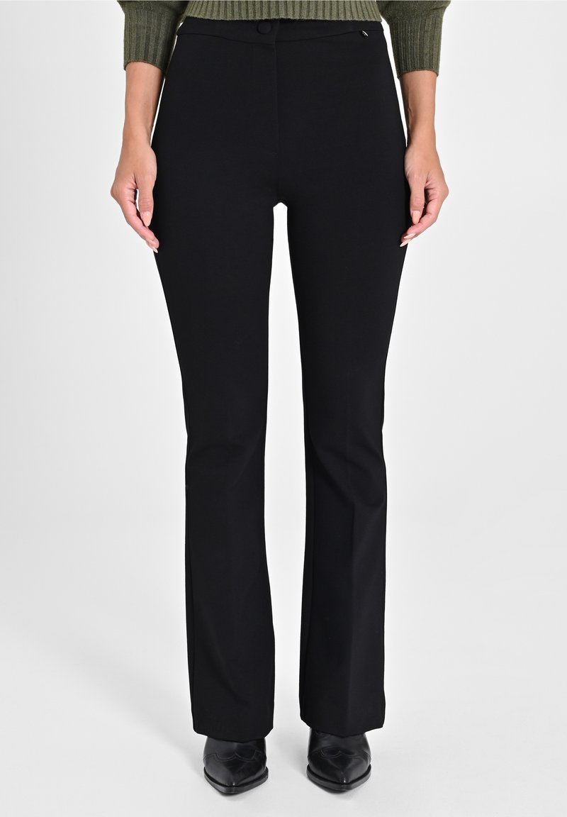 Femme portant un pantalon noir droit associé à des chaussures noires pointues et un pull côtelé vert olive, debout devant un fond uni.