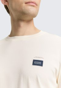 Kremowy t-shirt z krótkim rękawem i okrągłym dekoltem, ozdobiony niebieską łatą z tkaniny z napisem oraz przeszyciem nad nią. Miękka tekstura.