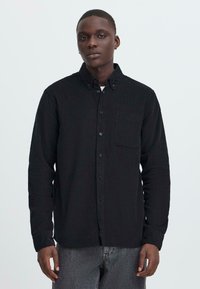 Black button-up shirt in textured fabric, featuring a pointed collar, front pocket, and long sleeves, worn over gray pants.