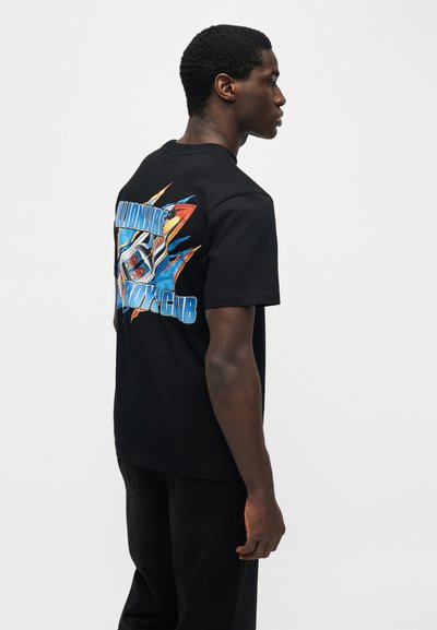 Άνδρας με μαύρο μπλουζάκι με πολύχρωμο γραφικό και το κείμενο «Billionaire Boys Club» στην πλάτη, στέκεται προφίλ μπροστά σε απλό φόντο.