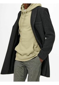 Lange grijze wollen jas over een lichtgroene hoodie met een voorvak, gecombineerd met geruite broek. Kenmerkt zich door een gestructureerde vorm en zachte texturen.