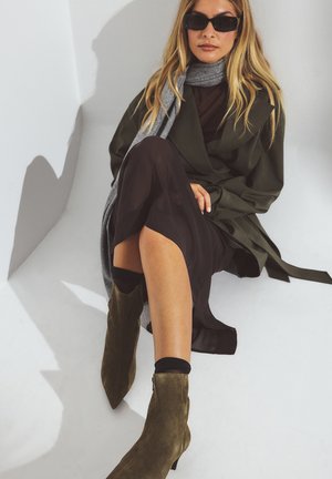 Abrigo trinchera verde con una bufanda gris, vestido negro y botas de tacón alto olivo. Texturas suaves y elementos de diseño a medida.