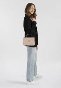 Rosa gesteppte Handtasche mit einer Kettenriemen, rechteckige Form, mit einem silbernen Dreieck-Logo auf der Vorderseite. Das Model trägt eine schwarze Jacke und helle Jeans.