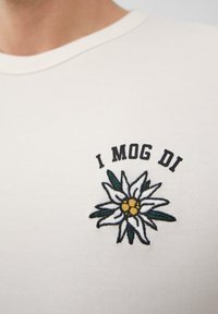 Weißes Baumwoll-T-Shirt mit einem gestickten Blumenmuster, das grüne Blätter und eine gelbe Mitte zeigt, begleitet von schwarzem Text "I MOG DI."