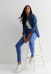 Jeansowa koszula narzucona na białą koszulkę bez rękawów, połączona z niebieskimi rurkami i białymi trampkami. Stylizacja charakteryzuje się swobodnym krojem i gładkimi fakturami.