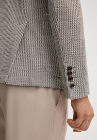 Braun-weißer gemusterter Blazer mit taillierter Passform, ausgestattet mit einem Reverskragen und vier braunen Knöpfen an den Ärmeln. Glatte Stofftextur.