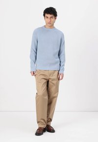 Pull en maille côtelée bleu clair avec un col rond, porté avec un pantalon beige ample et des chaussures marron foncé, se tenant devant un fond blanc.