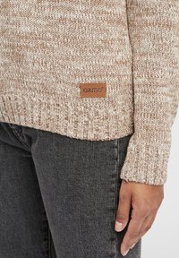 Neulepaita beige- ja kerma sävyissä ribbaussuutteilla ja helman. Sisältää ruskean nahka-patchin, jossa on "OXMO" logo vyötärön lähellä.