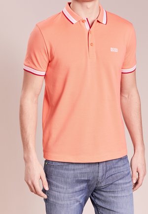 Mann trägt ein lachsfarbenes Poloshirt mit weißen und roten Besätzen an Ärmeln und Kragen, kombiniert mit hellblauen Jeans.