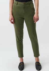 Pantaloni verde oliva slim fit, con chiusura a bottone, tasche frontali e una texture liscia. Abbinati a mocassini neri.