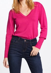 Fuchsia Langarmshirt mit V-Ausschnitt und taillierter Passform, kombiniert mit dunkelblauen Jeans im klassischen Fünf-Taschen-Stil und Kupferbeschlägen.