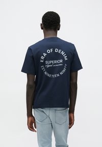 Μπλουζάκι από βαμβάκι σε ναυτικό μπλε με κοντά μανίκια· διαθέτει λευκό τυπωμένο κείμενο στην πλάτη: "ERA OF DENIM," "SUPERIOR," και "EST. NINETEEN NINETY."