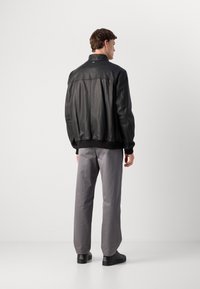Hombre vestido con una chaqueta de cuero negra, pantalones grises y zapatos negros, de pie dando la espalda contra un fondo blanco liso.