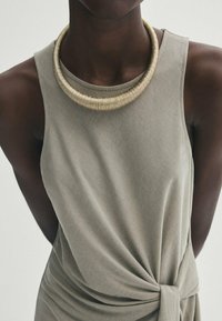 Collier en or avec des bandes texturées, porté sur un haut sans manches gris clair avec un détail de nœud sur le côté et un tissu doux.