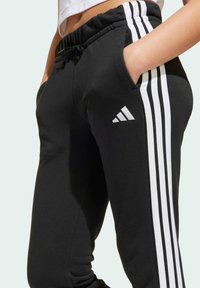 Čierne tepláky z mäkkej tkaniny so bielym trojitým pruhom na stranách a logom Adidas na pravej stehne. Elastický pás s vreckami.