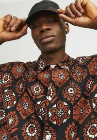 Camisa de manga corta con un fondo negro y patrones florales intrincados en marrón y blanco. Cuello abotonado y textura de tela ligera.