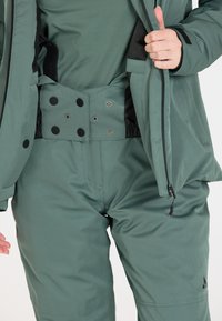 Grüne wasserdichte Jacke und Hose mit glatter Textur, ausgestattet mit Druckknöpfen, Reißverschlüssen und einer schmalen Passform. Logo auf der Hose sichtbar.