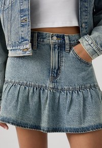 Φούστα από ανοιχτό μπλε τζιν με εφαρμοστή μέση, κλείσιμο με κουμπιά και βολάν στο τελείωμα. Διαθέτει ξεφτισμένες λεπτομέρειες και ελαφρώς ξεβαμμένη υφή.