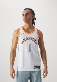 Camiseta de baloncesto blanca con detalles en naranja, que presenta "SAN ANTONIO" en letras marrones y el número "1". Material de malla transpirable, con aberturas laterales.