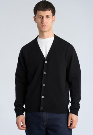 Junger Mann trägt eine schwarze, geknöpfte Strickjacke über einem weißen Hemd und blauen Jeans, steht vor einem schlichten hellgrauen Hintergrund.