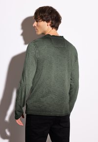 Zelená košile s dlouhým rukávem a texturovaným povrchem, s vlnitým výstřihem a volným střihem, v kombinaci s černými kalhotami.