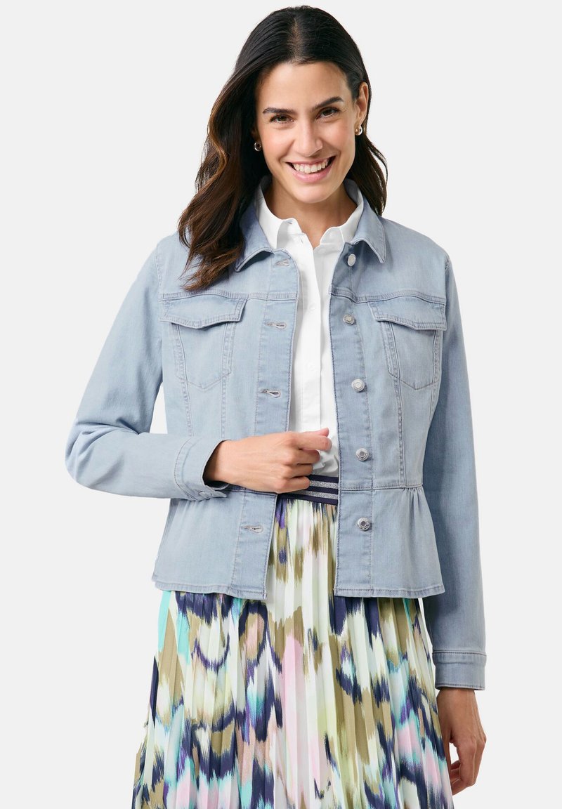 Hellblaue Jeansjacke mit Knopfleiste, zwei Brusttaschen und langen Ärmeln, kombiniert mit einem gefalteten, bunten Rock mit abstraktem Muster.