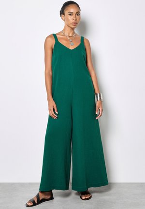 Donna che indossa una tuta verde senza maniche a gambe larghe, con collane d'argento sovrapposte, un ampio braccialetto d'argento e sandali neri, in piedi contro una parete bianca.