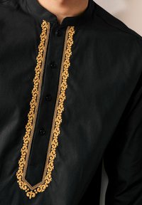 Chemise noire avec col montant, ornée d'une broderie dorée complexe le long de la patte de boutonnage avant et de quatre boutons noirs. Texture lisse.