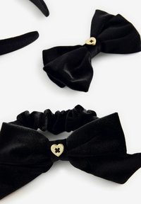 Gli accessori per capelli in velluto nero includono una fascia e un scrunchie, entrambi decorati con grandi fiocchi. Lo scrunchie presenta un accento a forma di cuore dorato.