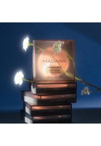 Compact en or rose pour le highlighter MÁDARA Moonlight Soft Glow avec un couvercle transparent, empilé sur des boîtiers noirs, entouré de petites fleurs blanches.