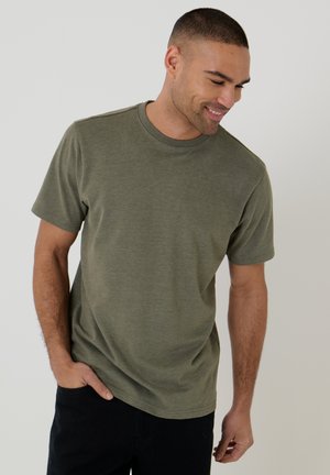 Uomo con i capelli corti sorridente, indossa una maglietta a maniche corte verde oliva e pantaloni neri, in piedi davanti a uno sfondo chiaro e uniforme.