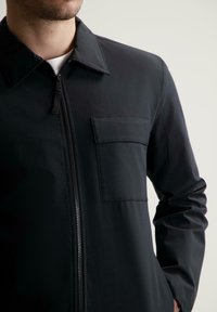 Giacca nera con zip in tessuto morbido, caratterizzata da un colletto, una chiusura frontale con zip e una tasca sul lato sinistro del petto. Il design è elegante e minimalista.