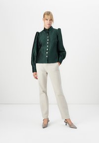 Grønn, teksturert bluse med høy hals, puffete ermer og en rekke skinnende, store knapper. Kombinert med lyse beige bukser og grå hæler.