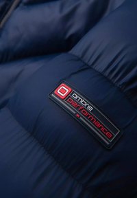 Sezione di una giacca imbottita blu navy con un'etichetta nera rettangolare con testo rosso e bianco, che recita "ombre performance." Texture liscia.