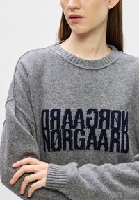 Pulôver cinza tricotado com tecido texturizado, gola redonda e mangas longas. Apresenta um logótipo azul escuro em destaque na parte frontal.