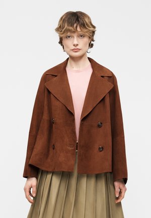 Persona con chaqueta de ante marrón de doble botonadura sobre una blusa rosa y falda plisada beige, de pie frente a un fondo claro y liso.