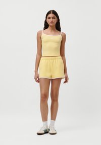 Gul tank topp med hvite stropper og matchende gule shorts med hvit kant. Modell iført hvite sokker og beige sneakers med grønne striper.
