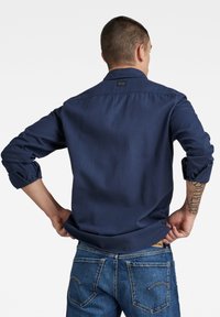 Chemise bleue marine à boutons avec manches retroussées, fabriquée en tissu lisse. Portée avec un jean en denim bleu avec des coutures contrastées et des poches arrière.