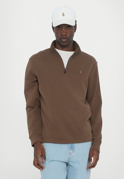 Lichtbruine rits-trui met een klein logo, gecombineerd met een witte baseballpet. Het model draagt ook lichtblauwe jeans. Gladde textuur, casual fit.