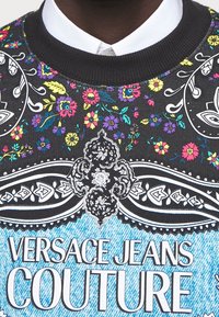 Tröja med svart tyg som har färgglada blommönster och paisleymönster. Inkluderar en sektion som liknar blå denim och texten "VERSACE JEANS COUTURE".