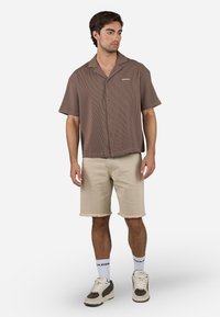 Brun kortærmet skjorte med knaplukning og vertikalt ribbet tekstur, parret med beige frayed denim shorts og sneakers i neutrale farver.