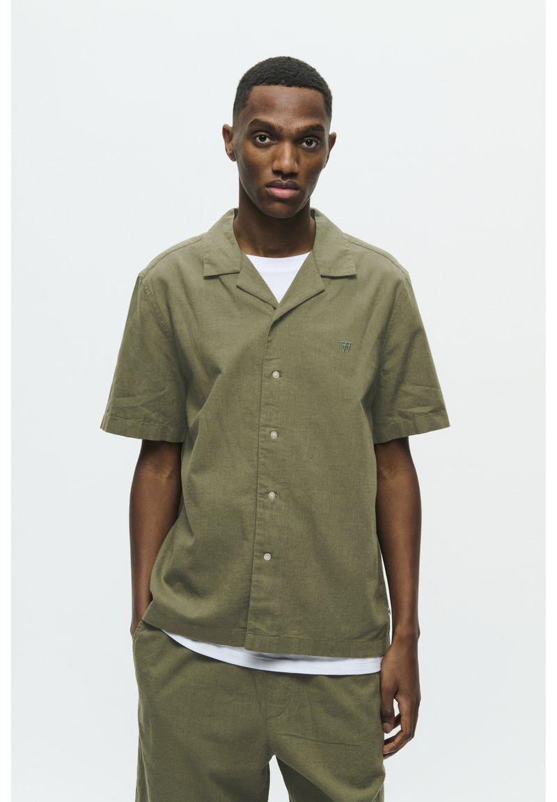 Chemise olive verte à manches courtes avec un col à encoches, des boutons sur le devant et un petit logo brodé sur le côté gauche.