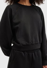 Czarna bluza z prążkowanym, okrągłym dekoltem, rękawami raglanowymi i subtelnymi, teksturowanymi detalami na ramionach. Elastyczny pas u dołu.