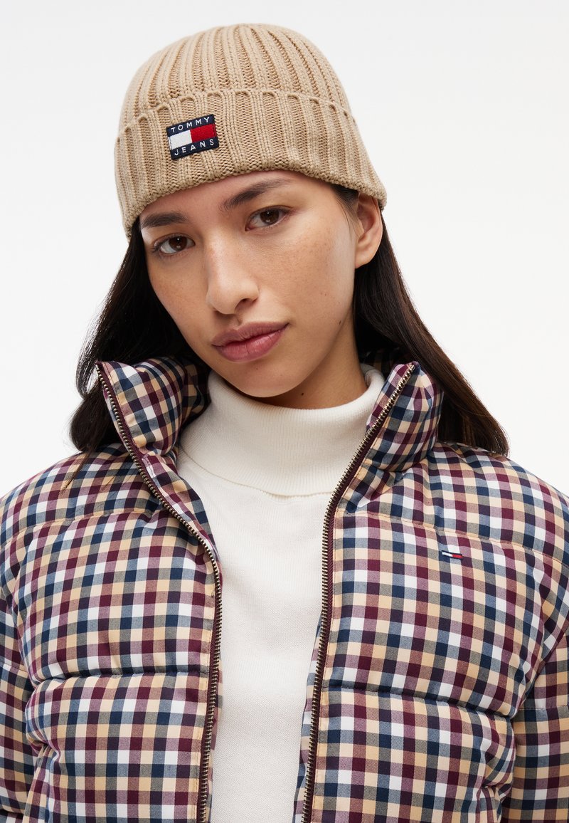 Femme portant un bonnet en tricot beige avec le logo Tommy Jeans et une doudoune à carreaux par-dessus un pull à col roulé blanc.