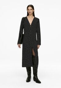 Schwarzes Polka-Dot-Kleid mit langen Ärmeln und tiefem V-Ausschnitt, mit einem seitlichen Schlitz und getragen mit kniehohen schwarzen Stiefeln.