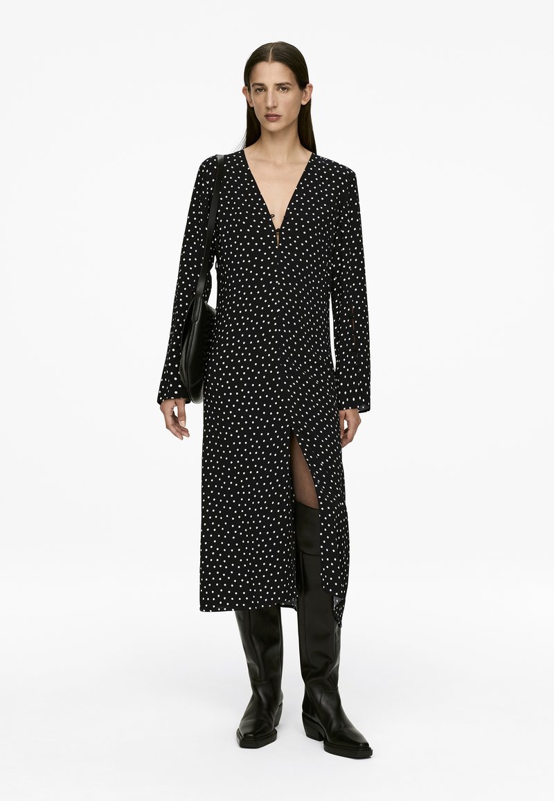 Schwarzes Polka-Dot-Kleid mit langen Ärmeln und tiefem V-Ausschnitt, mit einem seitlichen Schlitz und getragen mit kniehohen schwarzen Stiefeln.